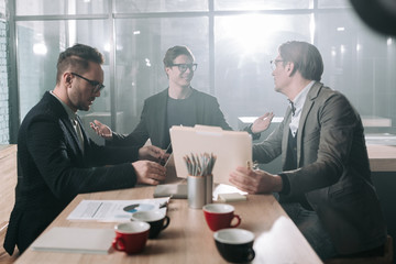 Aufkleber - Outgoing man flourishing arm while speaking with colleagues. They sitting at table. Serene adult watching at laptop. Business concept