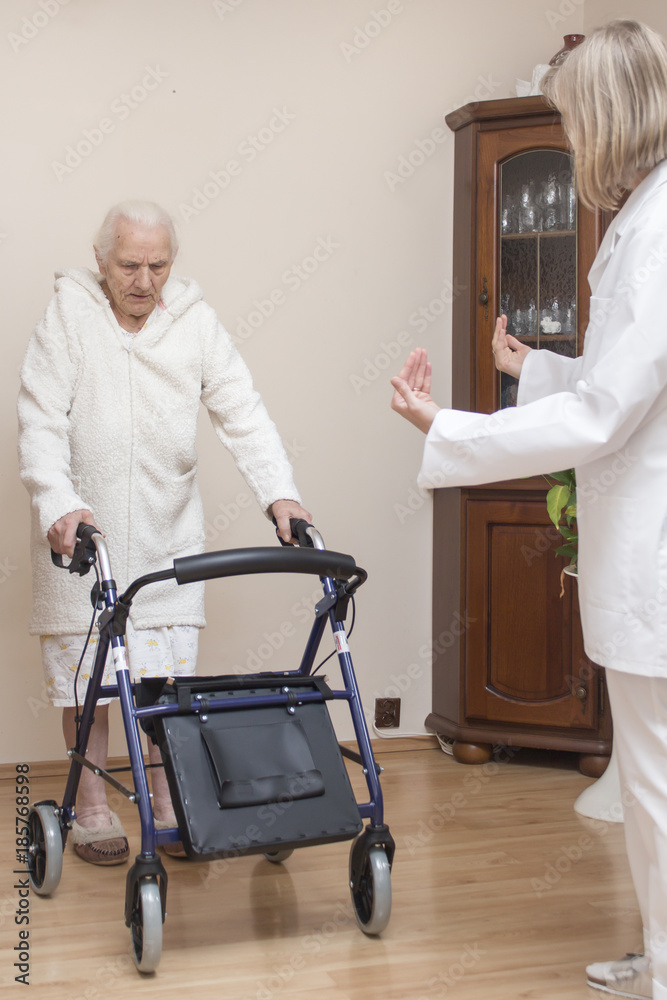 Staruszka w białym szlafroku uczy się chodzić z balkonikiem przy asyście pielęgniarki. - obrazy, fototapety, plakaty 
