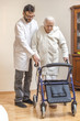 Staruszka w białym szlafroku chodzi przy pomocy chodzika przy asyście pielęgniarza.