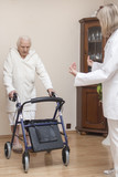 Fototapeta  - Staruszka w białym szlafroku uczy się chodzić z balkonikiem przy asyście pielęgniarki.