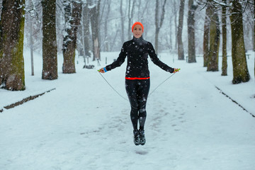 Beautiful sports girl doing exercises with jumping rope in winter park outdoor. Healthy lifestyle and cold weather concept.