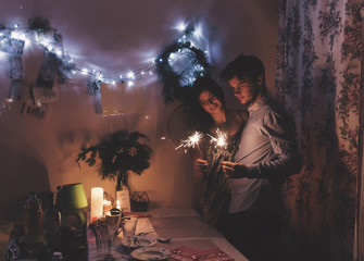 Wall Mural - happy stylish hipster couple holding burning sparklers fireworks and embracing in room at hoome in evening. happy holidays. happy new year and merry christmas concept. .