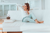 Fototapeta  - young woman in her late twenties sitting and laying on a lether white sofa in a cosy interier of her bright home and drinking a cup of tea and reading a book, totaly relaxed