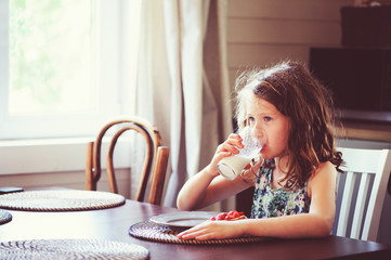 happy 8 years old child girl having breakfast in country kitchen, drinking milk and eating toast with strawberry