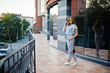 © AS Photo Family - Portrait of a perfect young woman wearing striped overall and yellow sunglasses poses with her handbag on a balcony of a modern building in a town.