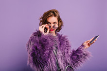 Interested Girl In Stylish Purple Fur Coat Looking To Camera Holding Sparkle Sunglasses. Indor Portrait Of Confident Female Model With Smartphone Posing On Bright Background.