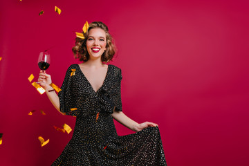 Blissful female model with shiny curly hair posing with wineglass on claret background. Emotional young woman wears black attire having fun at party with champagne and confetti and smiling.