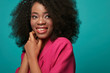 Studio portrait of attractive young african girl with afro hairstyle. Smiling girl wearing pink jacket, blue leggings and belt posing on turquoise  background