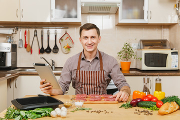 Young European man in apron sitting at table with vegetables, looking for recipe in tablet, cooking at home preparing meat stake from pork, beef or lamb, in light kitchen full of fancy kitchenware.