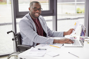 Elegant happy dark skinned male invalid looks over some paperwork, sit at work space next to big window, keyboards information on laptop computer. People, business, occupation, ethnicity concept