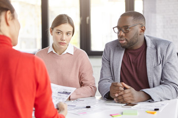 Dark skinned male and females coworkers sit together at working place, discuss opinions and ideas for new startup, plan strategies, have serious looks. Diverse crew of office workers have meeting