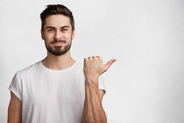 Wall Mural - Portrait of displeased bearded young male model wears white t shirt, indicates with thumb aside, sees something unpleasant, isolated over studio background with copy space for your advertisment