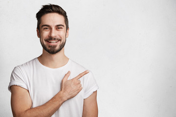 Horizontal portrait of bearded cheerful man has smile, wears casual white t shirt, indicates with fore finger at copy space for your promotional text or advertisment, isolated over concrete wall