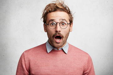 Wall Mural - Indoor shot of amazed shocked young man with mustache, stares through round spectacles, being surprised to hear unexcpected news, can`t believe eyes, isolated over white concrete background.