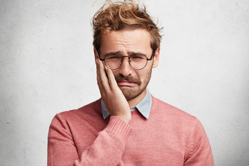 Wall Mural - Sorrorful bearded stylish guy has toothache, keeps hand on cheek, looks with desperate misreable expression, isolated over white concrete background. Handsome frustrated sad man feels sorry.