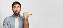 Horizontal Shot Of Amazed Young Man Has Thick Dark Beard, Dressed In Fashionable Clothes, Indicates With Thumb Aside, Shocked To Notice Something, Copy Space For Your Promotional Text Or Hearder