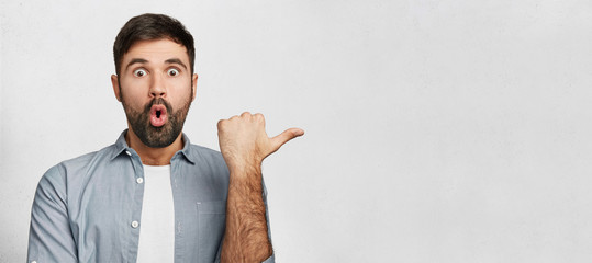 Wall Mural - Horizontal shot of amazed young man has thick dark beard, dressed in fashionable clothes, indicates with thumb aside, shocked to notice something, copy space for your promotional text or hearder