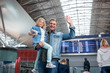 © Yakobchuk Olena - Our joyful trip. Low angle portrait of cheerful middle-aged father is holding his little daughter while standing at international airport and waving. They are looking at camera with wide smile
