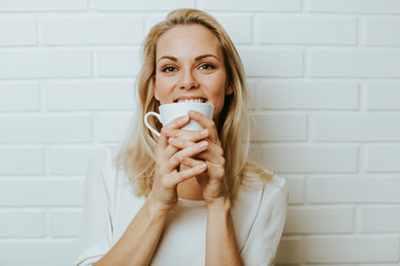 Wall Mural - Beautiful blond  caucasian woman posing in front of a white brick backround and  having fun with a cup or two cups of coffee, maybe tea