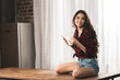 © LIGHTFIELD STUDIOS - smiling girl in checkered shirt and denim shorts using smartphone and drinking coffee at home