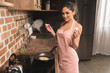 © LIGHTFIELD STUDIOS - sexy young woman in apron holding eggshell and smiling at camera while frying egg at kitchen