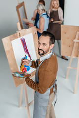 Sticker - handsome mature artist holding palette while standing near easel and smiling at camera