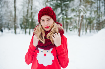 Blonde girl in red scarf, hat and santas sweater posing at park on winter day.