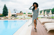 Attractive, pretty, tender, beautiful american woman in long dress in stripes and black hat. Model walking near pool of luxury hotel in Asia.