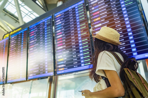Fotomural Young woman with backpack and carry on luggage in international airport, near flight information board