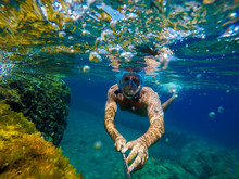 Underwater View Of A Young Diver Man Swimming In The Turquoise Sea Under The Surface With Snorkelling Mask For Summer Vacation While Taking A Selfie With A Stick.