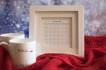 Full Winter Month Calendar January 2018 With Phases Of The Moon On A Blurred Background Of Snowy Window. The Calendar Is In The White Frame. Two Cups And A Red Knitted Scarf Are Next On The Table Too.