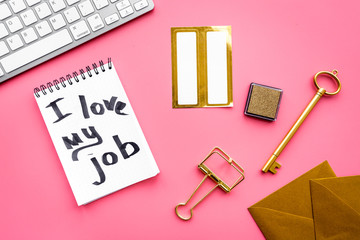 Happy at work. Handwritten motto I love my job in notebook near stationery on pink background top view