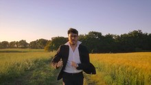 Running To The Future. Young Businessman With Elegant Suit Throwing Briefcase In The Middle Of Green Golden Wheat Field In The Nature At Sunset Steadycam Front View Dolly Slow Motion