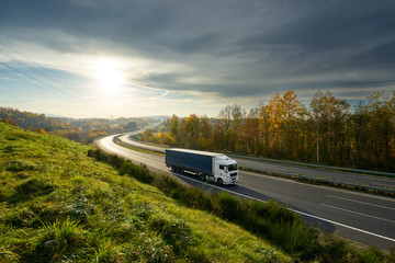 Poster - White truck driving on the highway turning towards the horizon in an autumn landscape with sun shining through the clouds in the sky