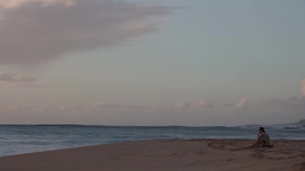 Wall Mural - a woman sitting on a beach sand watching ocean waves at sunset time