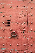 Closeup View Of An Old Red Door Of Medieval Style With Metallic Rivets, Heart Shaped Keyhole Plate, Cast Iron Knocker And Wire Mesh Spyhole.