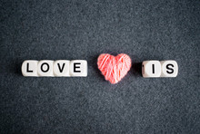 LOVE IS Text Word Made With White Plastic Blocks And Pink Thread Heart On Grey Felt Background. Love, Romance, Valentine's Day Concept