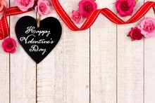 Happy Valentines Day Message On A Heart Shaped Chalkboard Tag With Ribbon And Flower Border Against A Rustic White Wood Background