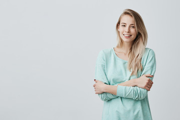 Wall Mural - Studio shot of pretty blonde girl with perfect teeth and healthy clean skin having rest indoors, smiling happily after receiving good positive news. Beautiful young woman standing with folded arms
