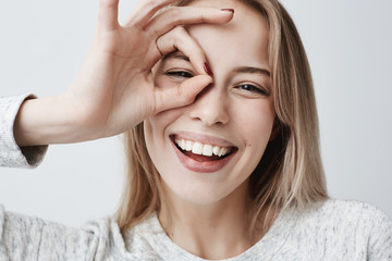 Wall Mural - Close up portrait of beautiful joyful blonde Caucasian female smiling, demonstrating white teeth, looking at the camera through fingers in okay gesture. Face expressions, emotions, and body language