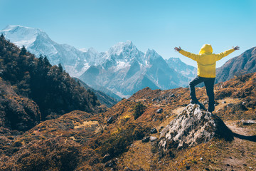 Wall Mural - Standing man with raised up arms on the stone and looking on snow covered mountains. Landscape with traveler, high rocks with snowy peaks, grass, trees in autumn in Nepal. Lifestyle, travel. Trekking