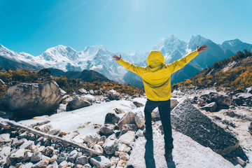 Wall Mural - Standing man with raised up arms near the river looking on snow covered mountains in bright day. Landscape with traveler, high rocks with snowy peaks, stones in autumn in Nepal. Travel. Trekking