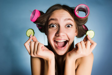 Wall Mural - the surprised young girl holds in hands pieces of a cucumber for humidifying a skin on the face, on a head of hair curlers