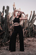 Vertical photo of a young female standing near big cactus in black trendy jumpsuit with eyes shut. Boho style. Bohemian, hippie concept