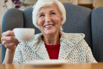 Poster - Head and shoulders portrait of cheerful elderly woman with charming smile looking away while enjoying delicious cappuccino at cozy small coffeehouse