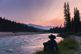 Fototapeta Do akwarium - Cowboy sitting by a raging river at sunset. The gorgeous colors of an Alberta sunset are displayed as the green grass grows and the river rushes by. The man sits with a denim jacket and cowboy hat.