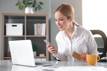 Poster - Portrait of glad businesswoman making purchase from internet. She watching at laptop and noting in phone. Job and e-commerce concept