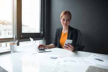 Aufkleber - Portrait of cheerful businesswoman noting in notebook computer while sending message in mobile. Technology and job concept
