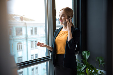 Poster - Portrait of happy businesswoman telling by mobile while standing near window in modern apartment. Conversation and labor concept