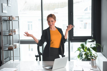 Poster - Portrait of glad businesswoman rising hands up while holding phone. She situating near desk with laptop in apartment. Occupation concept